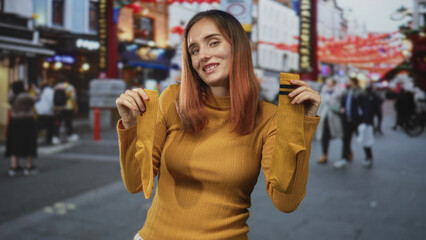 Fototapeta premium Young redhead woman holds mustard socks with raised arms on a busy city street, smiling and showing hosiery to camera; cozy playful.