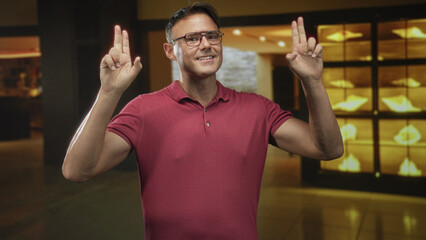 Naklejka premium Man raising hands with visible forearms in a building lobby showcase, wearing glasses and a red polo; casual confidence.