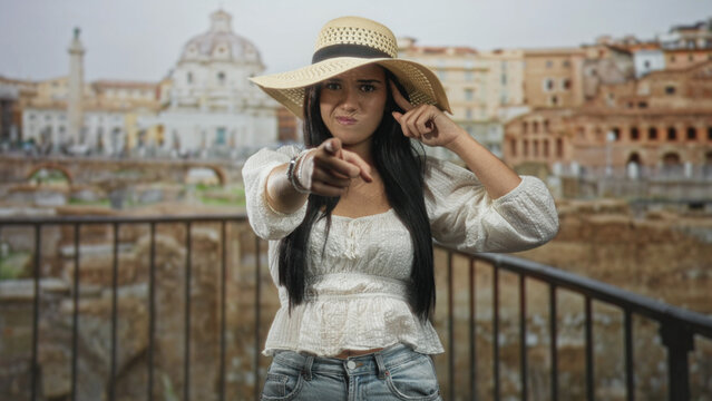 Woman points finger to camera with straw sun hat and bracelets standing by railing in front of ancient building ruins; blame disapproval.