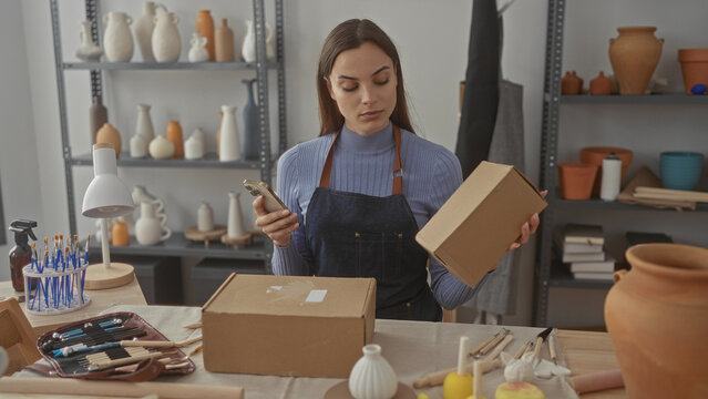 Woman wearing apron holds phone with one hand while touching cardboard box on workbench in artisan studio; concentration.