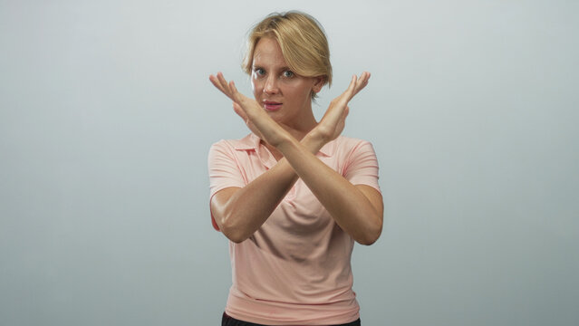 Blond woman in pink polo crosses forearms, hands forming an x over chest while gazing at camera in studio with plain backdrop; defiance.