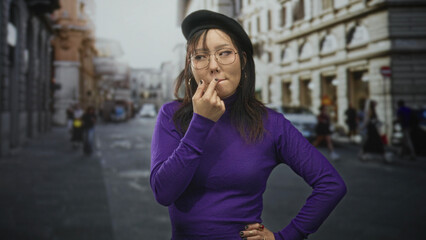 Fototapeta premium Young asian woman pinches her nose on a city street wearing purple turtleneck, beret and glasses; odor disgust.