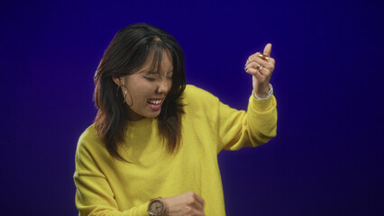 Woman making rock horns hand gesture with open mouth, hoop earrings, rings and nose piercing, yellow sweater in studio  playful joy. © Krakenimages.com