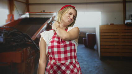 Woman cook in red check apron rubbing neck and grimacing inside a building with equipment and crates; discomfort.