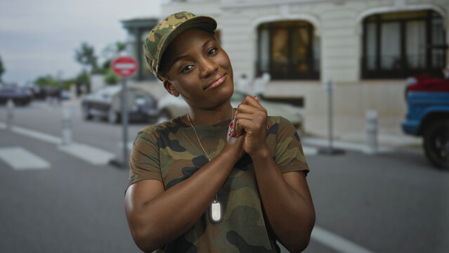 Woman in camouflage uniform clasps hands and dogtags while wearing a military hat on street near historic building; pride duty.