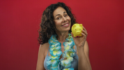 Woman wearing a lei and bikini joyfully holding a yellow piggy bank against a vibrant red background, showcasing a playful and tropical savings concept. © Krakenimages.com