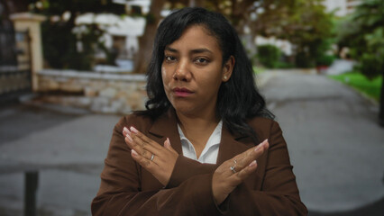 Fototapeta premium Woman in brown blazer stands with arms crossed in an x gesture on urban street with stone wall lining path; denial refusal rejection protest.