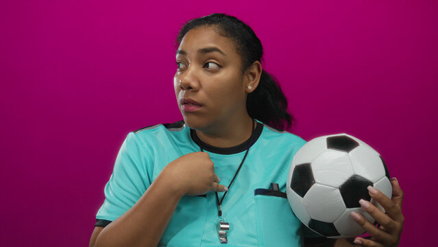 Woman referee wearing turquoise jersey pointing finger to chest and holding soccer ball in a pink studio; surprise.