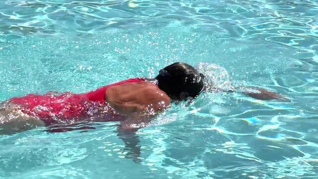 A middle aged woman swimming smoothly wearing a bright red bathing suit and goggles in a sparkling blue outdoor pool