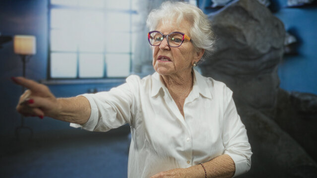 Elderly woman with crossed arms and pointing finger, wearing glasses and white shirt, standing by window and floor lamp in studio; stern disapproval.