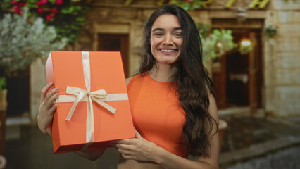 Woman holding large orange gift box and pointing finger at camera on street outside building entrance; celebration surprise.