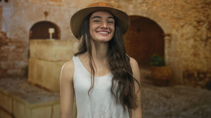 Fototapeta premium Teenage woman wearing straw hat and white tank top points finger to sunlit street in old town; adventure joy.