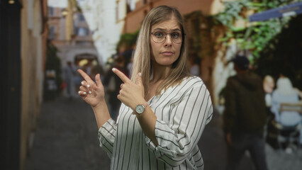 Fototapeta premium Blonde woman in glasses pointing finger down narrow cobblestone street beside outdoor cafe tables; uncertainty.