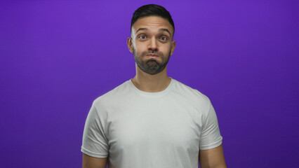 Young hispanic man playfully gesturing with a puffed face against a vibrant purple background wearing a plain white shirt displaying cheerful expressions.