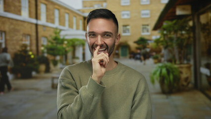 Naklejka premium Young man smiling playfully with finger over lips in a lively urban street setting capturing a candid youthful energy and modern city life outdoors.