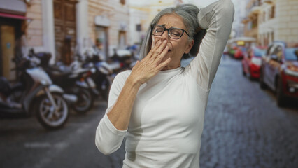 Fototapeta premium Middle aged woman holding neck with hands in narrow european street wearing glasses and white shirt, grimacing; pain.
