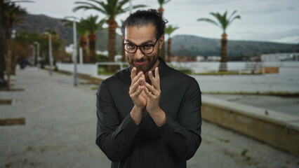 Man with glasses rubbing hands and pressing fingertips together on a street promenade by palm trees and waterfront; calm reflection.