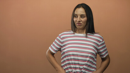 Obraz premium Woman grimacing in studio with peach background, wearing striped shirt and hands on hips showing a scrunched face; disgust discomfort.