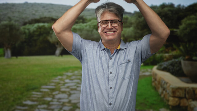 Man raises hands above head and smiles showing teeth on a stone path by a stonewall in forest wearing glasses and short sleeve shirt; joyful celebration.