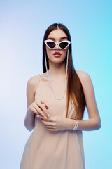 Fototapeta premium Young woman with long hair wearing white sunglasses and a beige dress poses confidently against a light blue background. Concept of beauty, fashion, and modern style.