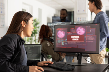 Asian female IT professional sits at desk staring at device screen displaying critical error...