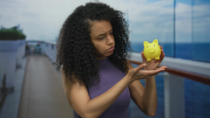 Woman holding yellow piggybank on cruise ship looks thoughtful against ocean backdrop, embodying travel and financial planning themes, with a curious seaside exploration vibe. © Krakenimages.com