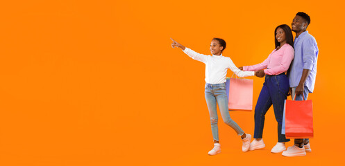 A girl is pointing at something while holding her mother's hand. Father stands nearby holding shopping bags. They have fun on a shopping day together with bright colors around them. © Prostock-studio