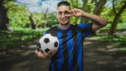 Fototapeta premium Man holding soccer ball with left hand and making a peace sign to his eye while wearing a blue striped jersey in forest path, smiling at camera; playful confidence.
