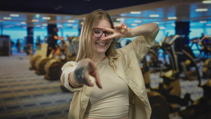 Fototapeta premium Blonde woman with glasses pointing finger at camera and making peace sign in gym building; playfulness optimism.