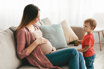 Young pregnant mother playing with her son, sitting on sofa and touching her belly, talking to her little child while spending time together at home