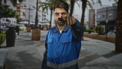 Man in blue work overalls points arm forward while looking down on a street, visible beard and reflective stripe; duty reliability.