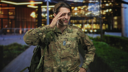 Obraz premium Man soldier in camouflage with backpack and dog tags, hand on forehead outside a lit building; duty fatigue.