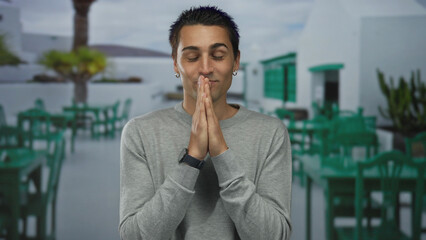Young man in gray sweater stands smiling with hands together on a sunny outdoor terrace with green chairs and tables in the background of a bright restaurant setting © Krakenimages.com