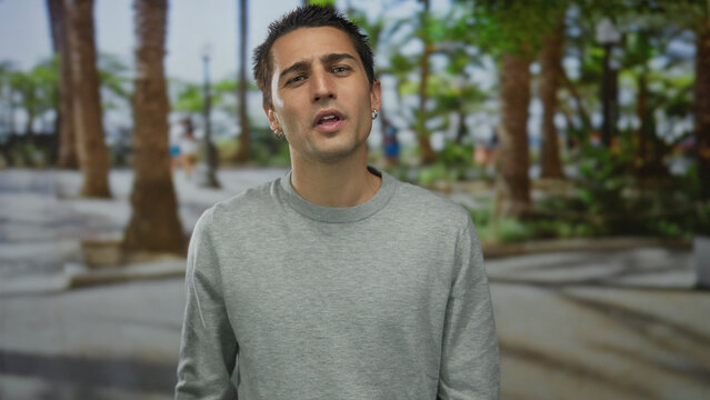 Young hispanic man flipping middle fingers in park amid palm trees with scowling expression; defiance.