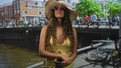 Hispanic teenage woman holding smartphone, wearing straw sunhat and pink sunglasses, looks up while...
