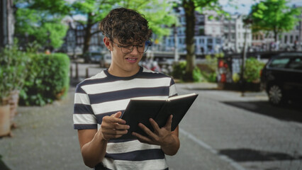 Fototapeta premium Teen boy in striped shirt holding open book with hands, reading on a tree lined urban street near parked cars; quiet concentration.