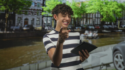 Fototapeta premium Boy in striped shirt points finger to tablet while holding the device on a street by a canal with parked cars and trees; playful curiosity.