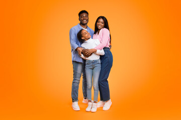 A family stands closely together with smiles on their faces. They are dressed casually. The background is bright orange. The scene shows warmth and happiness in their expressions. © Prostock-studio