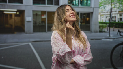 Fototapeta premium Woman covers mouth with hand while smiling on street near sidewalk in front of office building; playful shyness.
