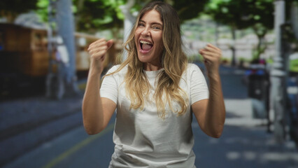 Fototapeta premium Middle aged woman raising clenched fists and smiling on a city street in a white t shirt; joy celebration triumph.