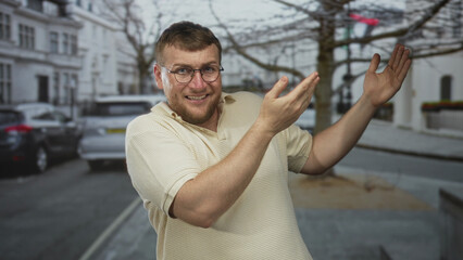 Fototapeta premium Man wearing glasses and a beige polo gesturing with both hands and open palms on a city street near parked cars and white buildings; surprise awkwardness.