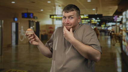 Man holding cupcake and hand covering mouth gesture in airport terminal near gate signage, looking...