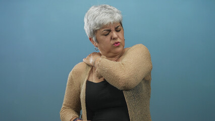 Woman with grey hair clutching her right shoulder, grimacing and wincing in a studio with a blue...