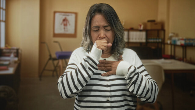 Hispanic middleaged woman coughing into fist with hand on chest in clinic building during a medical exam; illness concern.