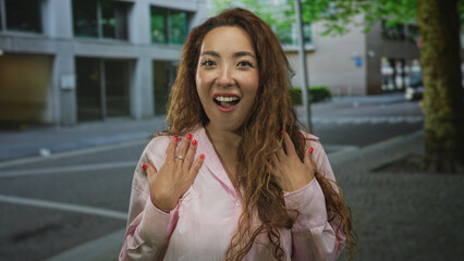 Fototapeta premium Woman with hands to chest and mouth open in surprise on a city street near building and tree, pink blouse and red nails; surprise reaction.
