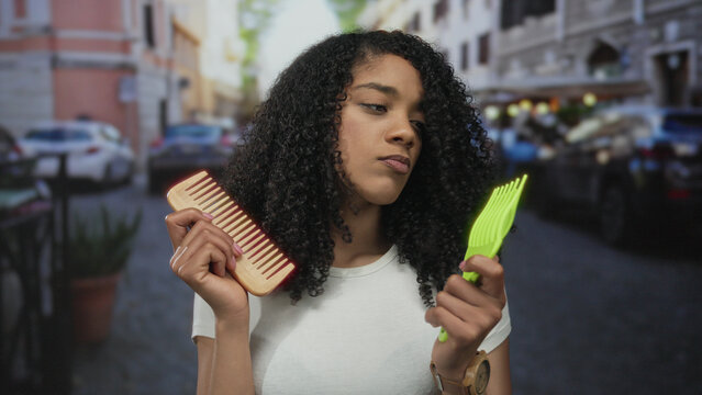 African american woman holds wooden comb and green pick as she compares hair tools on bustling street; indecision.