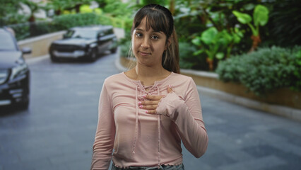 Young woman with raised hand and other hand on chest on street, eyes closed in pledge or apology...