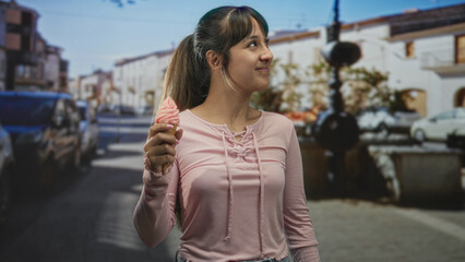 Obraz premium Young woman holding swirled strawberry ice cream cone, smiling and looking up while standing on a sunny street lined with cars and buildings; sweet contentment.
