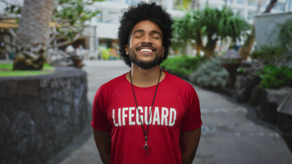 Smiling man in a red lifeguard shirt stands outdoors in a city street with greenery. © Krakenimages.com