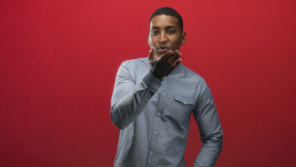 Obraz premium Young black man with hand to lips blows a kiss, covers mouth and points with fingers in a red studio; affection connection.